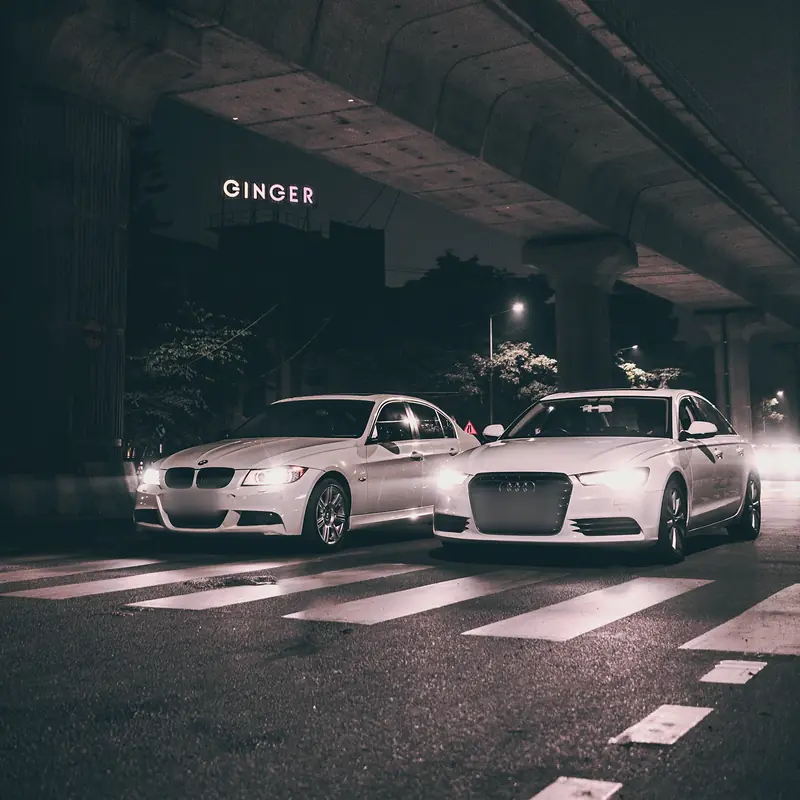 Two white saloons, headlights on, under a concrete viaduct at night